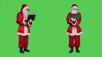Santa uses laptop on greenscreen, wearing festive christmas costume with white beard and browsing website. Modern saint nick works on portable pc, online network in studio. photo