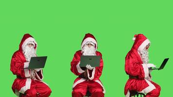 Young man as santa claus using laptop, posing dressed in red and white festive suit during christmas holiday season. Saint nick sitting on chair scrolling on website, xmas celebration. photo