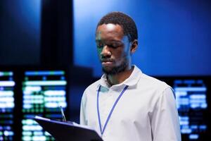 Engineer doing yearly maintenance in order to extend data center hardware life span and avoid damages. African american specialist monitoring server infrastructure components photo