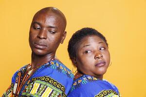 Black man and woman looking at each other while standing back to back studio closeup portrait. Confident african american couple with serious facial expression posing on orange background photo