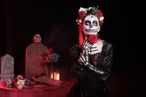 Woman using red rose for magic ritual in graveyard, celebrating Day of the Dead. Mysterious lady acting as occult witch, surrounded by frightening funeral items during Halloween horror event photo