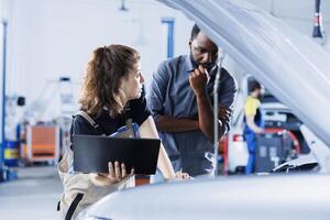 Mechanic in car service uses laptop to calculate invoice after fixing broken automobile battery. Skillful garage worker uses device to inform woman of final costs after mending vehicle photo