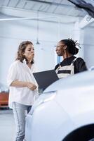 African american mechanic in car service uses laptop to calculate invoice after fixing broken automobile engine. Garage worker uses device to inform woman of final costs after mending vehicle photo