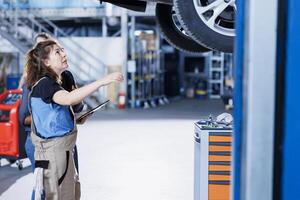 Experienced mechanic in garage using laptop to follow checklist while doing maintenance on car, talking with client. Specialist in auto repair shop does checkup on vehicle wheels next to customer photo