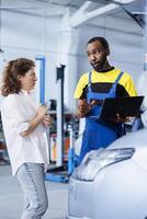 Experienced mechanic in car service orders new engine for damaged vehicle using laptop. Adept engineer next to customer looking online for components to replace old ones in malfunctioning automobile photo