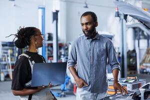 BIPOC woman next to client in car service orders new engine for damaged vehicle using laptop. Garage employee looking for components on internet to replace old ones in faulty automobile photo