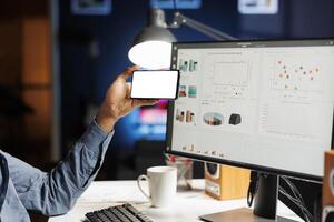 Freelancer examines white screen on phone and analysis on pc, uses multiple gadgets at workstation. Male contractor looking at blank display layout, calculating cost and revenue elements. photo