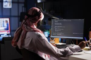 Islamic man shows how to design an app while sitting at desk and using a computer. Over-the-shoulder image of a pc monitor displaying active algorithms created by a traditional-dressed Arab guy. photo
