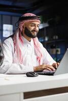 While seated at his home office desk, a Middle Eastern male entrepreneur works effectively on his laptop. Portrait of Arab man checking e-mail, perusing the internet, and thoroughly examining notes. photo