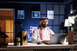 Arab businessman typing on his laptop at a trendy workstation. In a peaceful and professional workplace atmosphere, a Muslim man is browsing, communicating via email, and using WiFi connectivity. photo