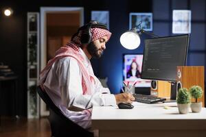 Middle Eastern software developer demonstrating the fundamentals of software development by entering code on a laptop. Arab man running algorithms while using wireless headphones. photo