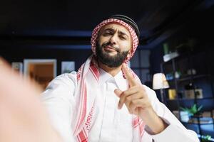 POV of a jovial Muslim man participating in a conference. Middle Eastern guy wearing traditional clothing is vlogging online while holding a smart gadget and pointing at the camera. photo