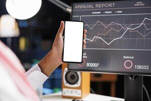 A close-up of a guy hand holding a mobile device with a white copyspace screen in front of a computer monitor. On a smartphone, an Arab individual displays a blank chromakey mockup template. photo