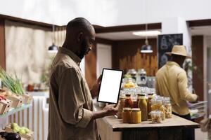 African American man checking the eco friendly products, jars, and groceries on shelves while holding a tablet with a chromakey template. Blank screens for mockups. Sustainable local supermarket. photo