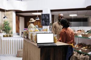 Placed on a wooden counter is a digital laptop showing an isolated white screen display. A wireless computer at a contemporary grocery shop has a blank chromakey mockup template. photo