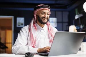 Enthusiastic Middle Eastern businessman efficiently works on his laptop while seated at his home office desk. Profile shot of Arab guy checking e-mail, surfing the net, and taking detailed notes. photo