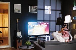 Muslim guy competes in a virtual reality game by using a wireless controller and showing distress by holding his head after losing. The monitor displays the detailed digital world. photo