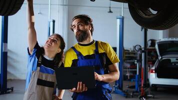 Knowledgeable mechanic and customer standing underneath car in garage using laptop to order new parts after finding malfunctions. Expert assisting client by looking for components over the internet photo