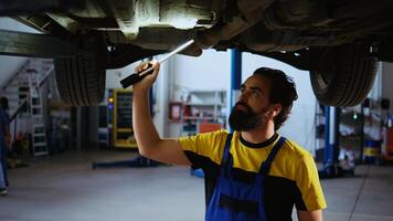Precise serviceman working on suspended car in garage, checking parts during routine maintenance. Expert in auto repair shop walking underneath vehicle, scanning it using work light photo