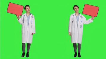 Woman doctor holding a red empty speech bubble sign in studio, showing an isolated blank cardboard icon against greenscreen backdrop. Physician creating announcement with banner. Camera A. photo