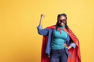 African american girl portraying superhero with red cape, isolated over studio background, flexing muscles. Smiling teenager wearing cloak posing as hero in costume, having fun photo
