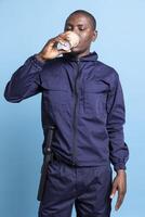 African american agent drinking coffee cup against blue background, serving caffeine refreshment to get more energy after a sleepless night. Male security guard poses in front of camera. photo