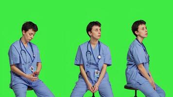 Medical assistant waiting for someone while she sits on chair, feeling impatient and waits for patients to attend checkup appointments. Nurse in scrubs against greenscreen backdrop. Camera B. photo