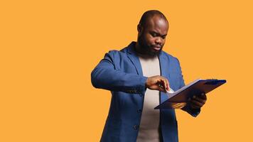 Man browsing though financial audit report on clipboard, scratching head, feeling puzzled, studio background. Stressed african american worker confused by paperwork on notepad, camera B photo