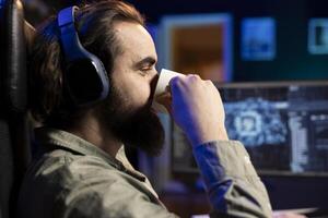 IT specialist drinking coffee with headphones on listening to songs, scripting artificial intelligence on computer. Developer enjoying hot beverage and music in ears while working on coding AI on PC photo