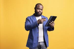 Irked man upset after lacking necessary money while trying to buy stuff online. Grumpy person looking at debit card after getting transaction rejected on tablet, isolated over studio background photo