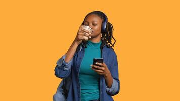 Woman scrolling on smartphone, using phone to check social media feed, drinking coffee. Girl browsing internet websites, enjoying beverage, listening music, isolated over studio background, camera A photo