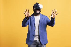 Joyous african american man wearing virtual reality headset, interacting with interface. Delighted person having fun using high tech futuristic modern VR gadget, isolated over yellow studio background photo