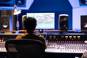 African american audio technician recording tracks from sound booth, processing tracks and working with digital software in control room. Expert pressing buttons and sliders for high quality. photo