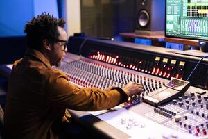 African american tracking engineer operating daw software to edit songs and control desk board to mix and master the tunes. Professional record producer creating new music, twisting knobs. photo