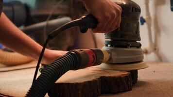 Craftsperson works with orbital sander on lumber block to achieve smooth finish, close up shot. Woodworking specialist in furniture assembly shop using angle grinder to fix wooden surface, camera A photo