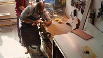 Sculptor shaping raw timber using chisel and hammer in carpentry shop with coworker cleaning workbench in background. BIPOC man making wood sculptures, engraving plank with tools, camera B photo