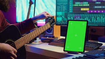 Musical producer learning to tune his guitar before playing, watching lesson tutorial by using greenscreen display on tablet. Young artist tuning strings on instrument, practicing. Camera A. photo