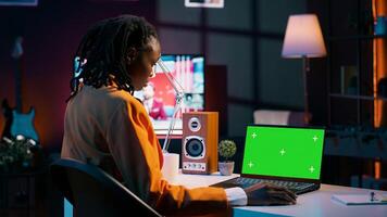 African american young woman looks at laptop with greenscreen, sitting at her home desk and preparing to attend online university lessons. Girl using pc with isolated mockup layout. Camera A. photo