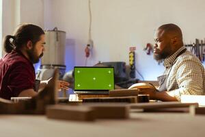 BIPOC artisan and apprentice comparing wood art piece with schematic displayed on green screen laptop. Woodworking specialists in studio crosschecking wooden object with mockup notebook blueprint photo