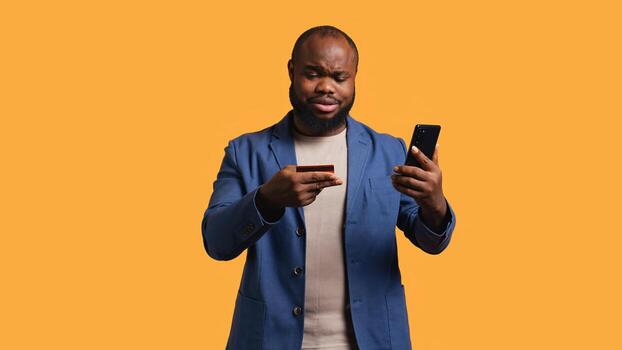 Man upset after lacking necessary money while trying to buy stuff online on smartphone. Sad person looking at debit card after getting transaction rejected on phone, studio background, camera B photo