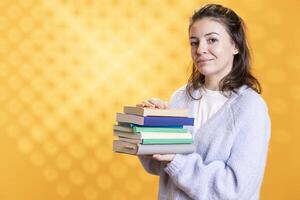 Portrait of merry woman smiling while holding pile of books, enjoying reading activity. Radiant person pleased with stack of novels in arms, enjoying leisure time, studio background photo