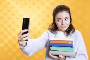 Smiling woman taking phone selfie with stack of books containing information for school exam, studio background. Student taking pictures with smartphone before university assessment, holding textbooks photo