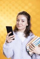 Portrait of smiling woman with stack of textbooks, gathering information for school exam, checking phone screen, studio background. Jolly person holding pile of books, reading messages on smartphone photo