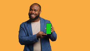 Smiling influencer presenting green screen mobile phone, isolated over studio background. Joyful BIPOC person holding copy space chroma key smartphone used for advertising brands, camera A photo