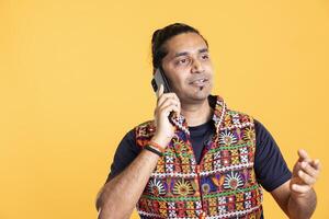 Smiling man chatting with friend over telephone call, enjoying relaxation time. Radiant person wearing traditional indian attire talking with mate in phone call, studio background photo
