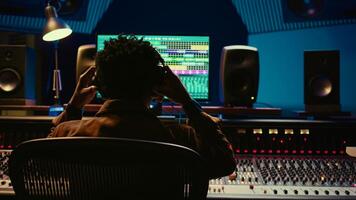 Professional tracking engineer using headphones to edit recorded tracks in control room, motorized sound control knobs. Audio expert working on mixing console and editing software. Camera B. photo