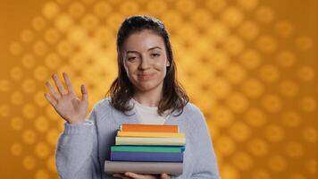 Portrait of smiling woman holding stack of books, doing salutation hand gesture, feeling optimistic. Jolly person with pile of novels raising arm to greet someone, studio background, camera B photo