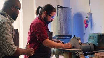 Man teaching african american trainee how to shape wooden bowl on disc sander in assembling shop. Worker learning from woodworking professional how to use grinding machine rotating disc, camera A photo