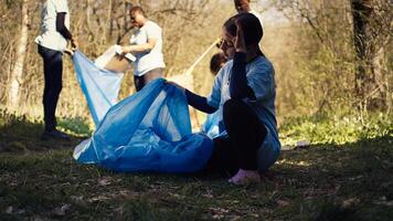Tired little girl collecting trash and plastic bottles from the forest, helping the volunteers with litter cleanup. Child activist picking up rubbish in a garbage disposal bag. Close up. Camera B. photo