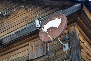 Old rusty satellite disk on wooden house with brown plank walls building corner photo